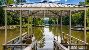 blue-bayou-dock-1 | Swack Vacations Wooden dock structure with a metal roof extends over a calm lake, surrounded by green trees and boats moored along the shoreline.