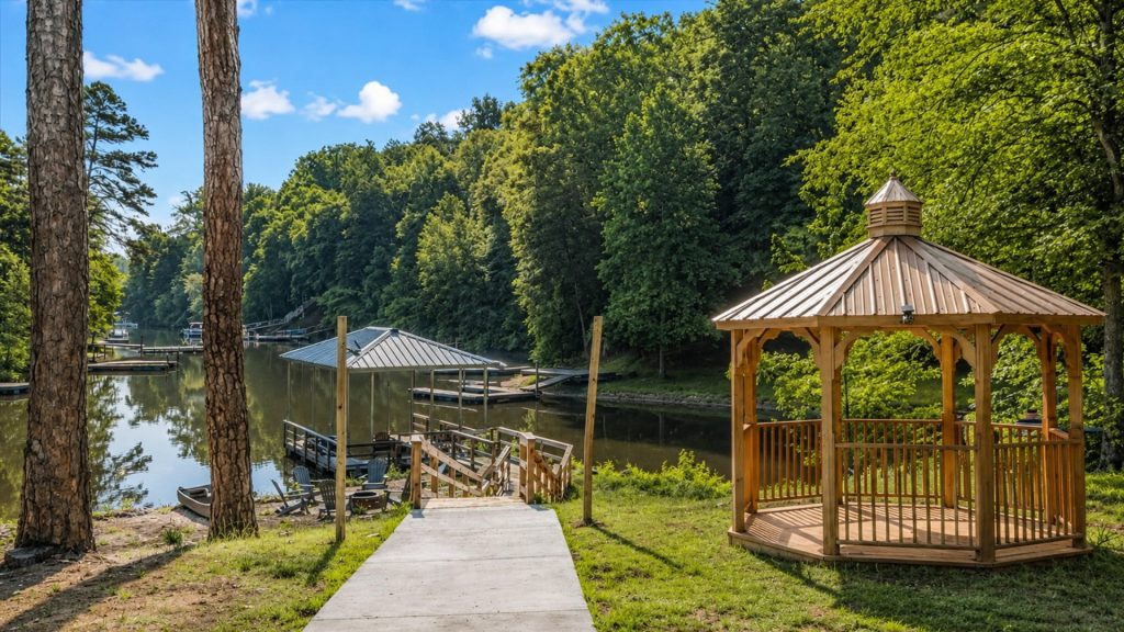 Wooden gazebo by a calm lake with docks and a concrete path, surrounded by tall trees under a blue sky.