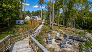 Wooden boardwalk leading to a cluster of white houses with a gazebo, surrounded by tall trees and blue sky.