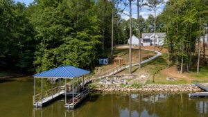 Lakeside residential yard with a blue-roofed dock, gazebo, winding path, and houses among tall trees by the water.