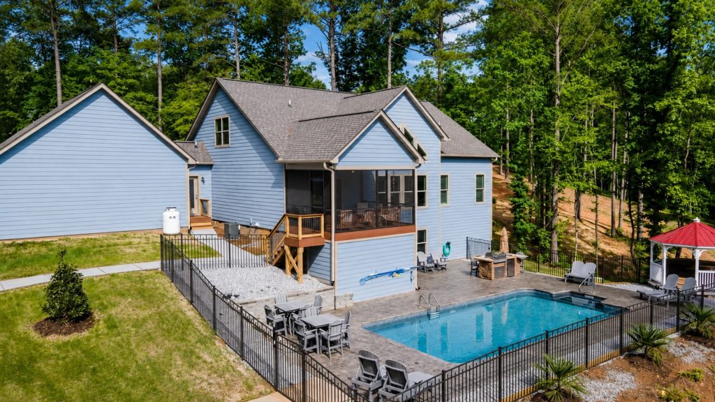 Blue two-story house with a gray roof, screened porch, and a fenced rectangular pool in a backyard surrounded by trees and greenery.