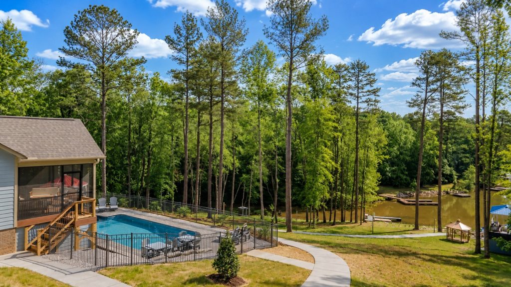 Backyard with a rectangular pool, screened porch, and wooden deck, surrounded by tall pine trees and a bright sky.