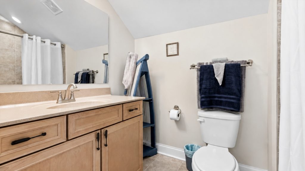Bathroom with light wood vanity, beige countertop, and large mirror, towels on a wall rack, and a toilet nearby.