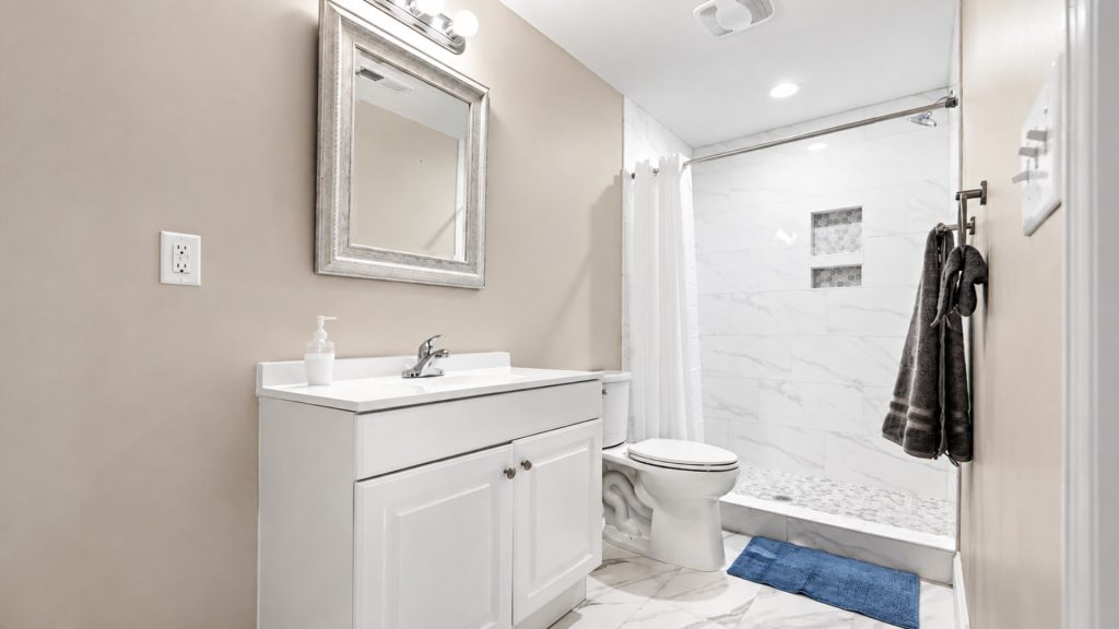 White bathroom with a vanity, framed mirror, and sink; a toilet beside it and a marble-tiled shower area with a towel rack on the wall. Two blue bath mats on the floor.