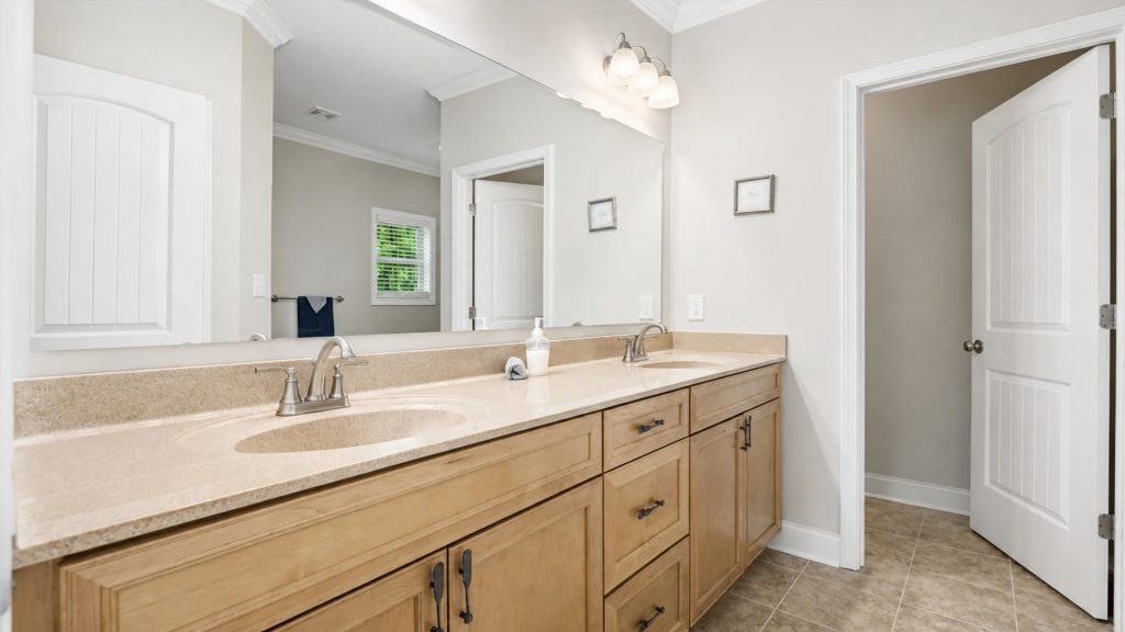 Double-sink bathroom vanity with beige countertop, wood cabinets, and a large mirror. A doorway is open to the right.