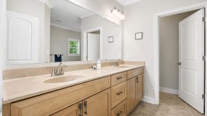 blue-bayou-full-bath-4-1 | Swack Vacations Double-sink bathroom vanity with beige countertop, wood cabinets, and a large mirror. A doorway is open to the right.