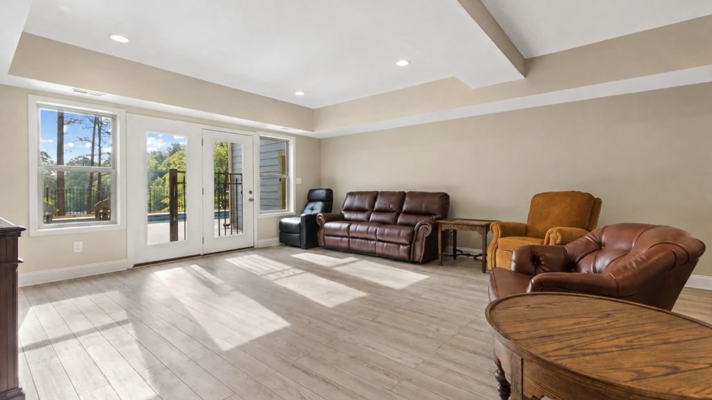 Sunlit living room with large glass doors to a patio and pool, beige walls, light wood floors, and brown leather seating surrounding a round wooden table.
