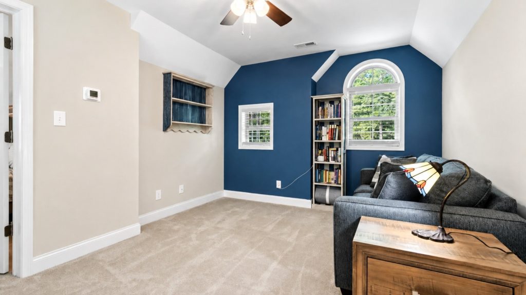Cozy living area with a dark blue accent wall, arched window, tall bookshelf, and gray sofa with a lamp and pillows.