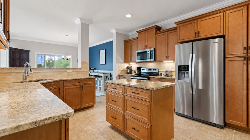 Bright kitchen with wooden cabinets, granite countertops, and a central island next to a stainless steel refrigerator.