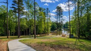 Curving concrete path through a sunlit forest toward a lake with docks and a gazebo under a blue sky with white clouds.