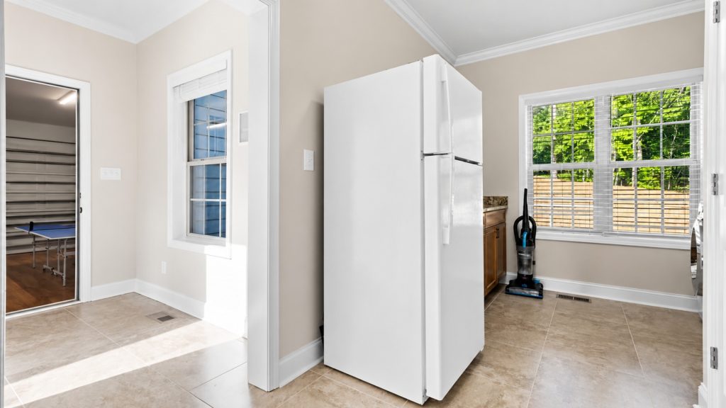 Bright kitchen area with a white, side-by-side refrigerator, beige walls, and a large window with blinds; a vacuum sits by the cabinet. The adjacent doorway reveals a room with a ping-pong table far left.