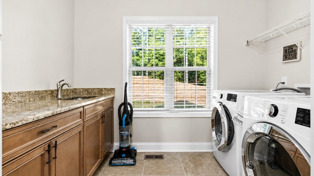 Laundry room with a granite countertop, sink, wooden cabinets, and a window with blinds beside a front-loading washer and dryer.