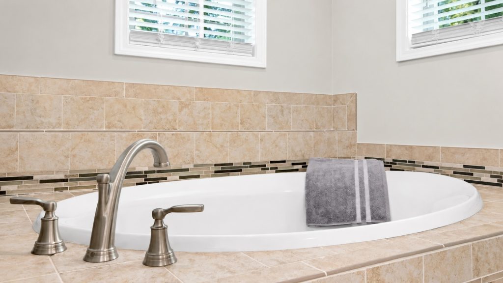 Oval white bathtub with brushed-nickel faucet and a gray striped towel draped over the edge, beige tile surround and two high windows above.