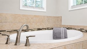 Oval white bathtub with brushed-nickel faucet and a gray striped towel draped over the edge, beige tile surround and two high windows above.
