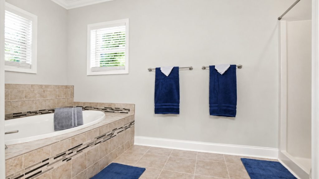 Bathroom with a tiled tub surround, two navy towels hanging on a chrome rod, and matching blue bath mats on beige tile floor.