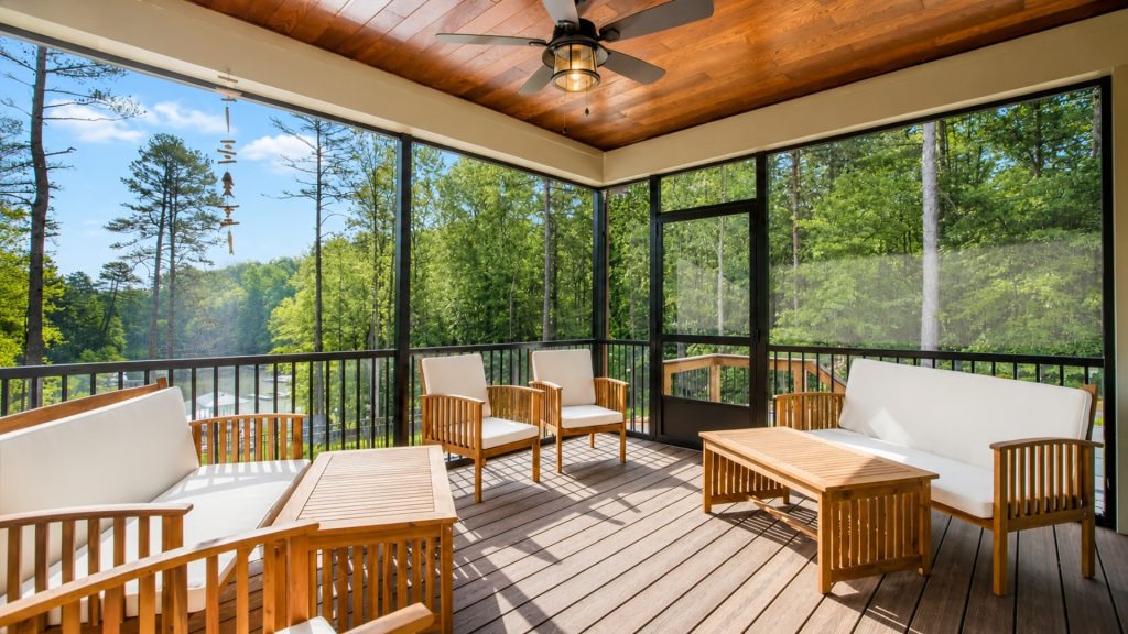 Screened wooden porch with white-cushioned seating and matching coffee tables, overlooking a green forest and trees outside.