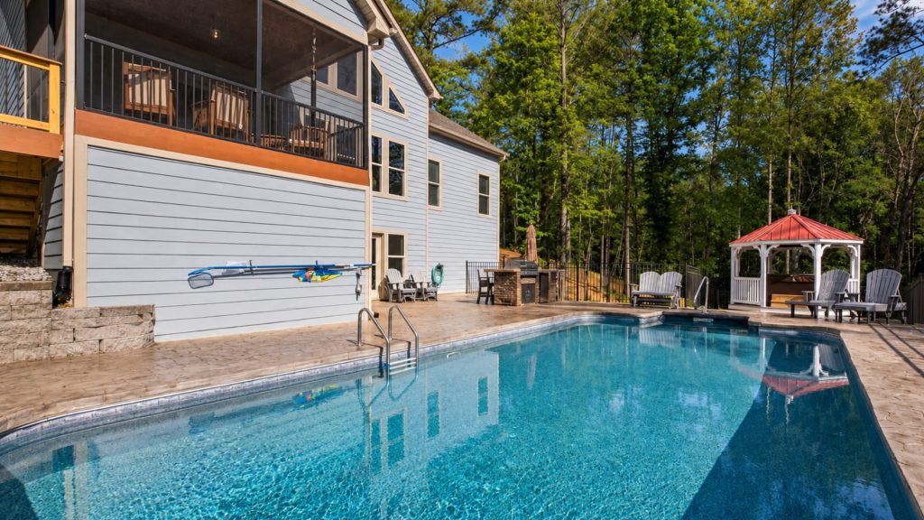 Backyard pool area with a light blue rectangular pool beside a gray house and a red-roof gazebo in the background.