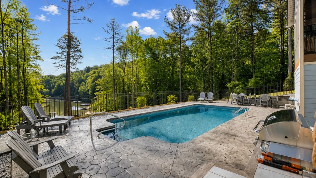 Poolside patio with gray hexagonal paving, Adirondack chairs, and a fence overlooking a forested lake.