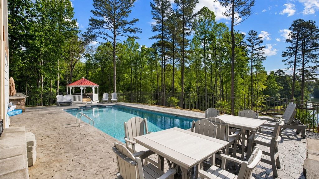 Poolside patio with white tables and chairs, a red-roof gazebo, and tall trees overlooking a lake on a sunny day, surrounded by a fence.