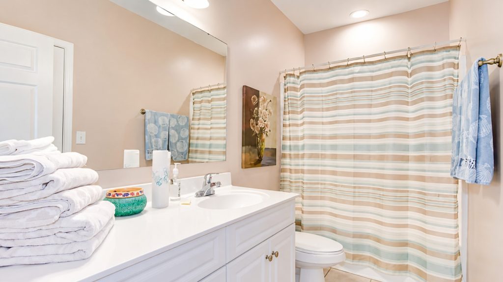 White bathroom vanity with folded towels, soap dispenser, and a decorative bowl on the counter; pale pink walls and a striped shower curtain in beige, blue, and white.