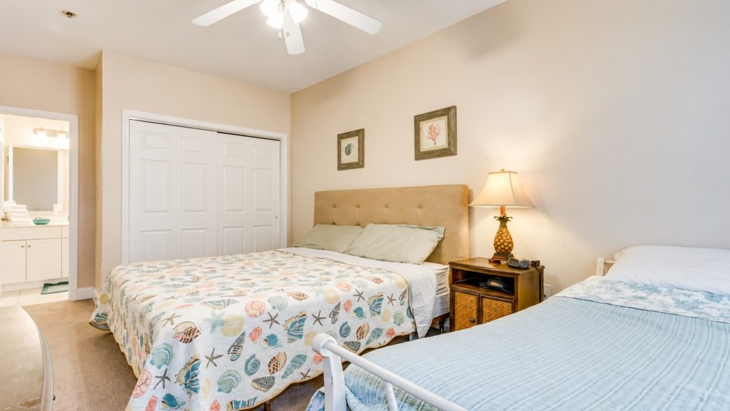 Two double beds in a bright hotel room with a seashell-pattern quilt and a wooden nightstand with a pineapple lamp beside them.