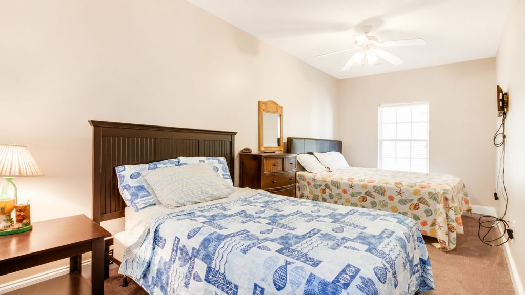 Bright hotel room with two beds, coastal-print quilts, a wooden dresser with a mirror, and a ceiling fan.