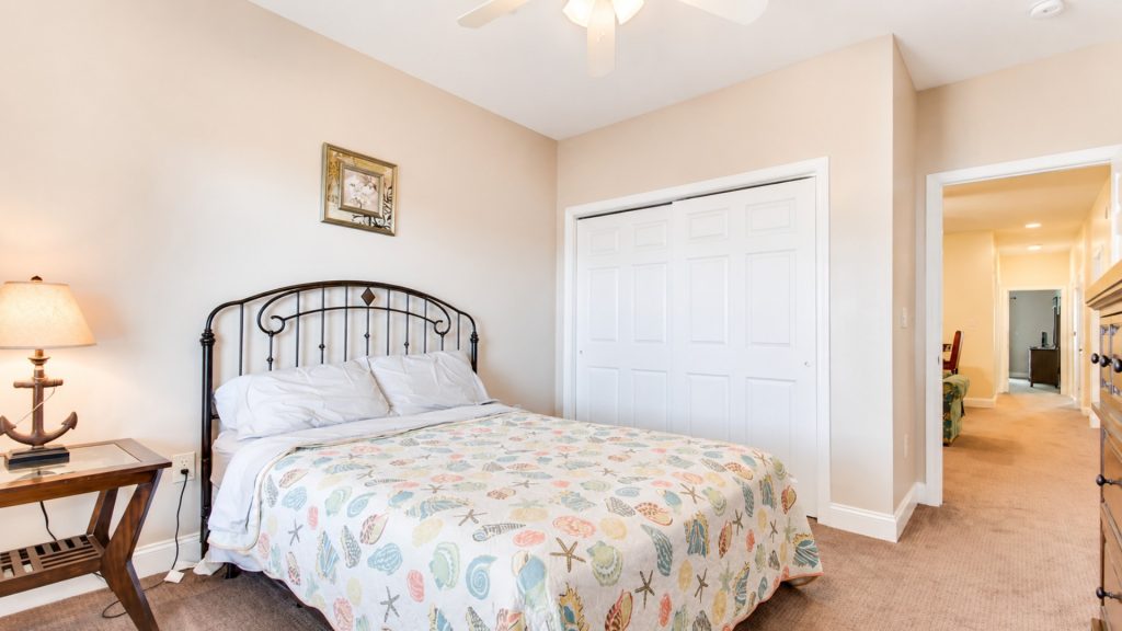 Bedroom with a metal headboard bed, seashell-patterned quilt, and a wooden nightstand with a lamp on beige walls. Closet doors to the right and a hallway visible in the background.]','Bedroom scene: bed, framed art, closet doors, and a bright hallway visible beyond.