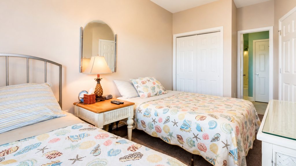 Twin beds with seashell-patterned quilts in a bright hotel room, with a nightstand and lamp between them.