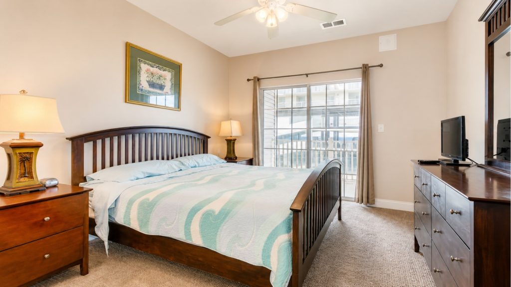 Bedroom with a dark wood bed, matching nightstands, and a turquoise patterned quilt; sunlight from a sliding glass door to a balcony fills the room.