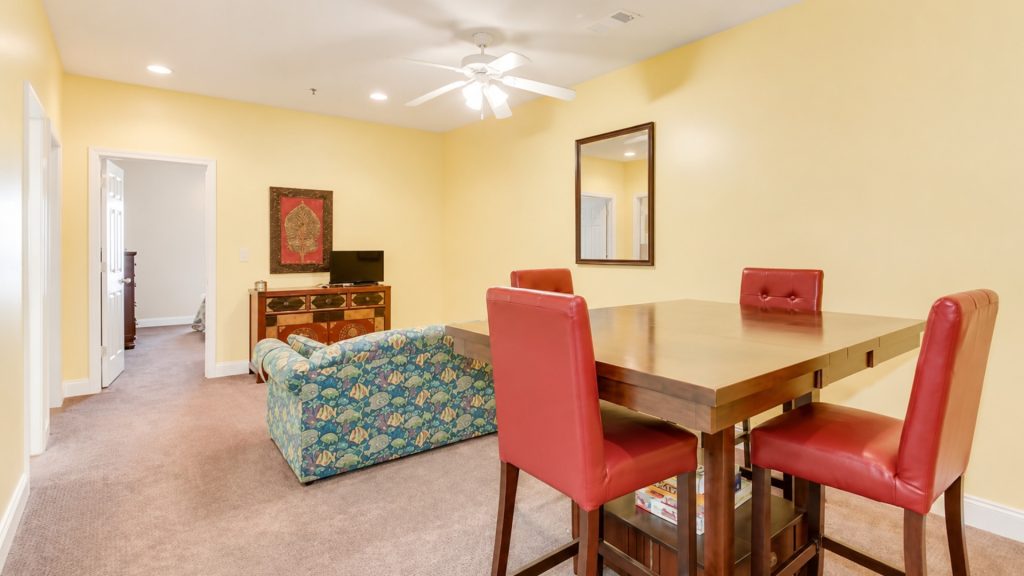 Bright dining and living area with a wooden table surrounded by four red upholstered chairs; yellow walls, patterned sofa, and a TV on a wooden cabinet nearby.