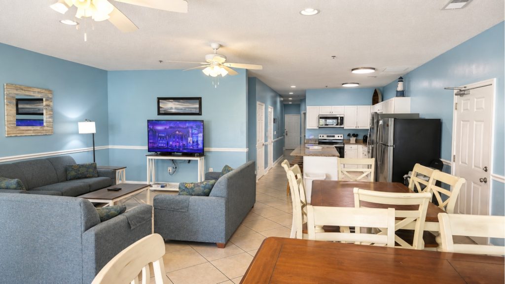 Open-plan living area with blue walls, gray sofas, and a TV against the wall, with a white-kitchen backdrop in view.