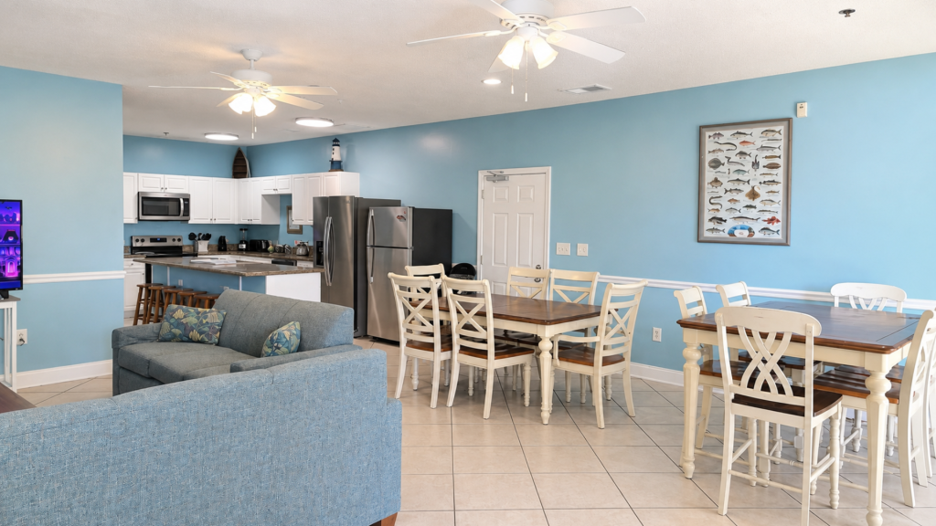 Open-plan living space with blue walls: white kitchen cabinets, stainless steel fridge, island with stools, and a wooden dining table.