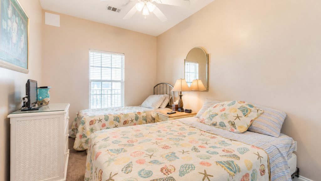 Twin bedroom with seashell-pattern bedspreads, matching pillows, and a nightstand between beds with two lamps.