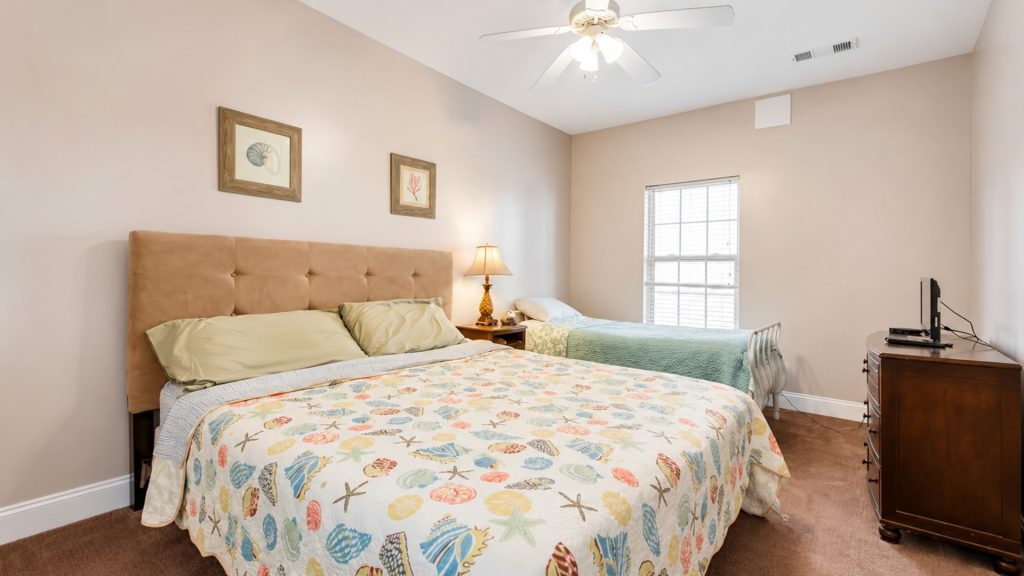 Two double beds with a beige tufted headboard and a seashell-themed quilt, a wooden nightstand with lamp between them, and a window to the right in a beige bedroom.