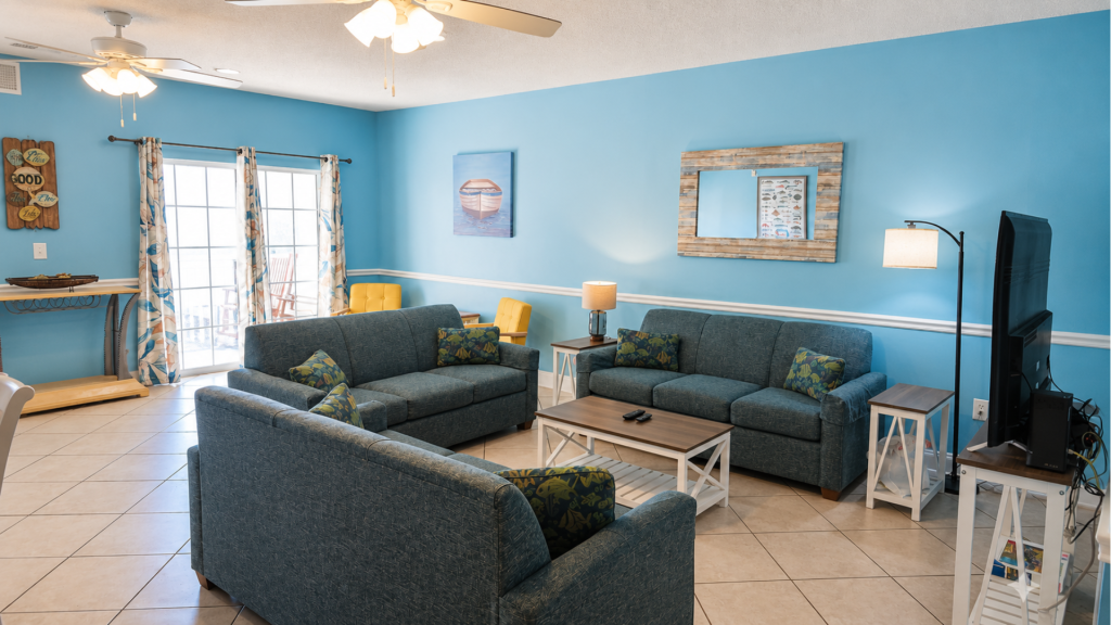 Coastal living room with blue sofas, tropical pillows, a wooden coffee table, and a TV on the right.