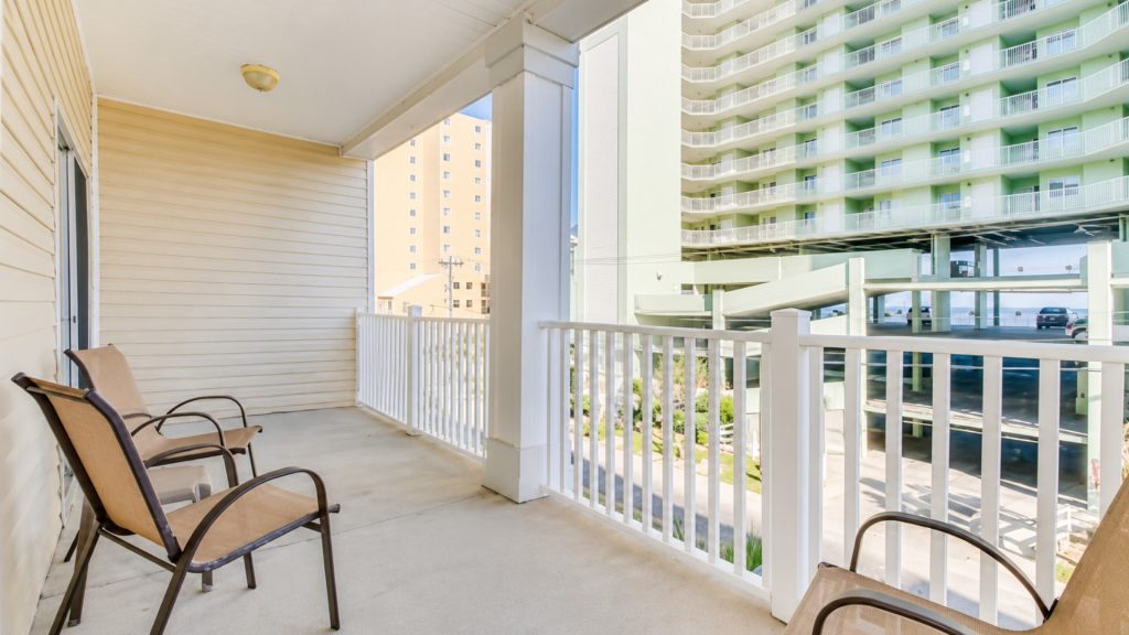 Balcony with beige siding and two tan chairs, white railing, overlooking a green apartment building and a parking structure.