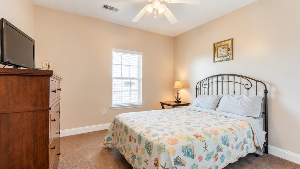 Bedroom with a metal headboard bed covered by a seashell-pattern quilt, against beige walls, a window, and a wooden dresser with a TV on top