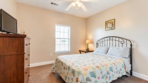 Bedroom with a metal headboard bed covered by a seashell-pattern quilt, against beige walls, a window, and a wooden dresser with a TV on top