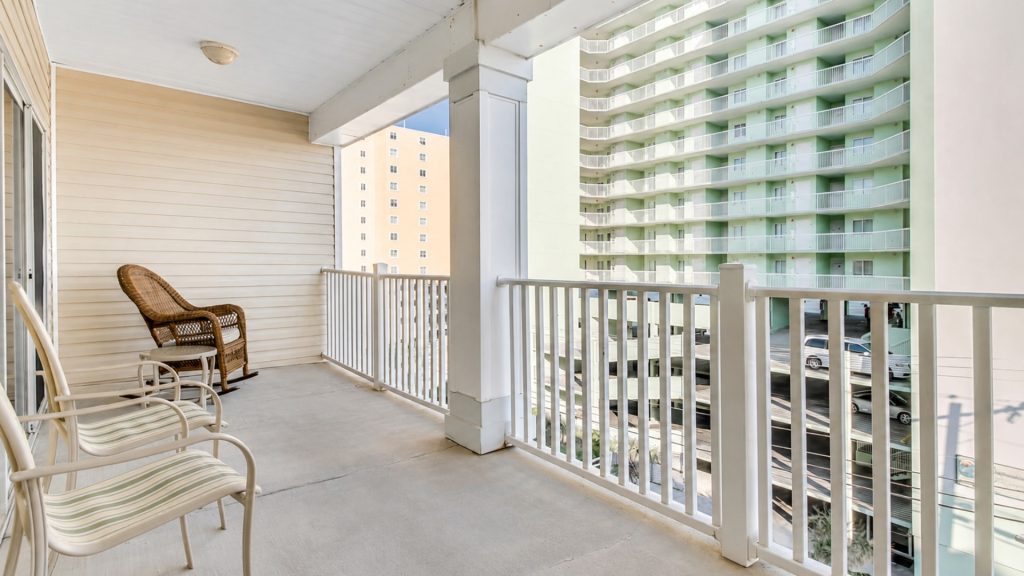 Balcony with white railing and seating: wicker chair and striped lounge chairs, overlooking a pale-green high-rise and a beige building in the background