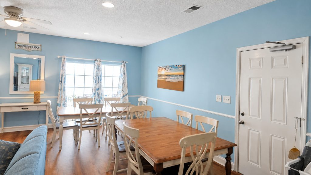 Coastal dining room with blue walls, wooden tables, and white chairs; window with patterned curtains and beach-themed wall art.