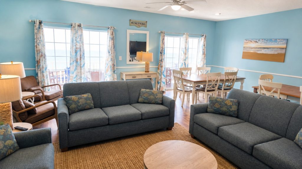 Coastal living room with blue sofas, patterned cushions, and a woven rug facing a round wooden coffee table.