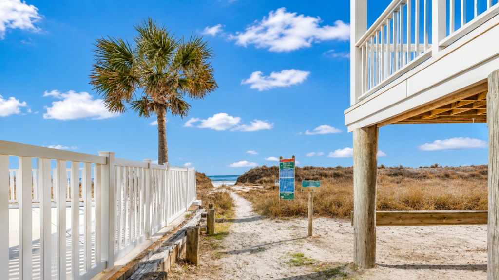 Beach path between white railings and a palm tree, under a bright blue sky with scattered clouds.