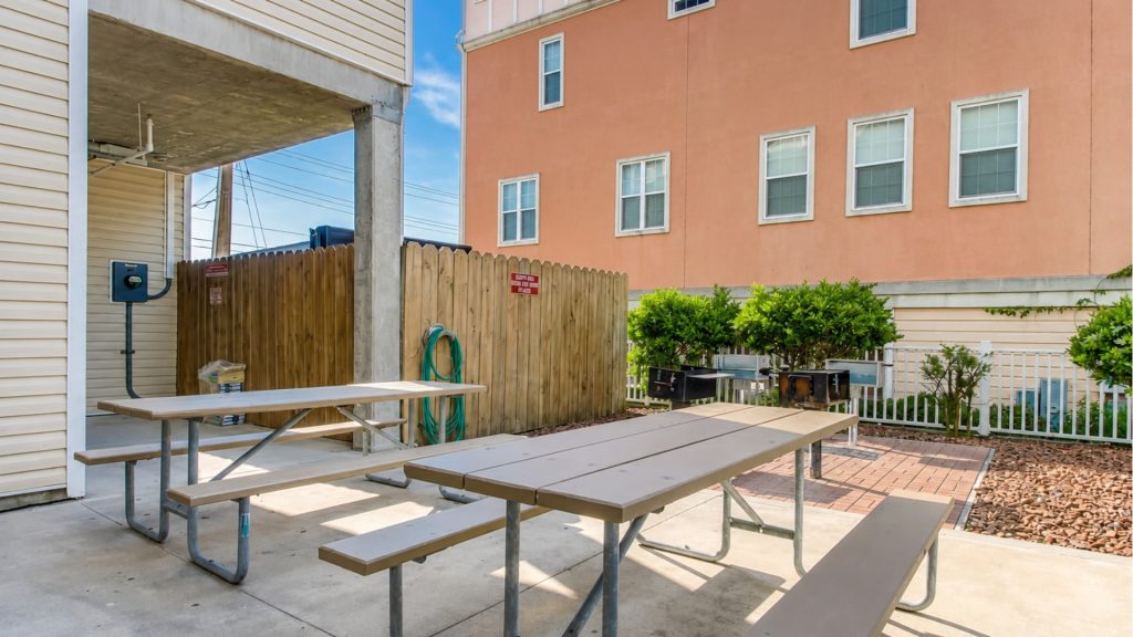Outdoor communal patio with two brown picnic tables on concrete, a wooden privacy fence, and green shrubs in front of a peach building.
