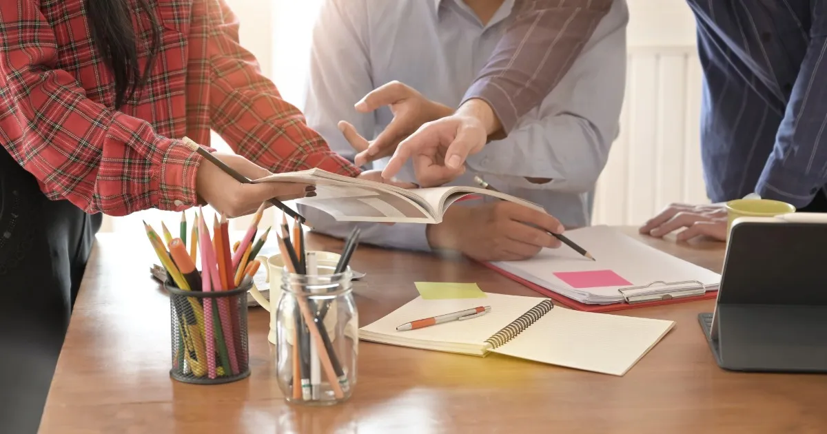 Group of people collaborating around a wooden table, with an open book and notebooks visible on the desk.