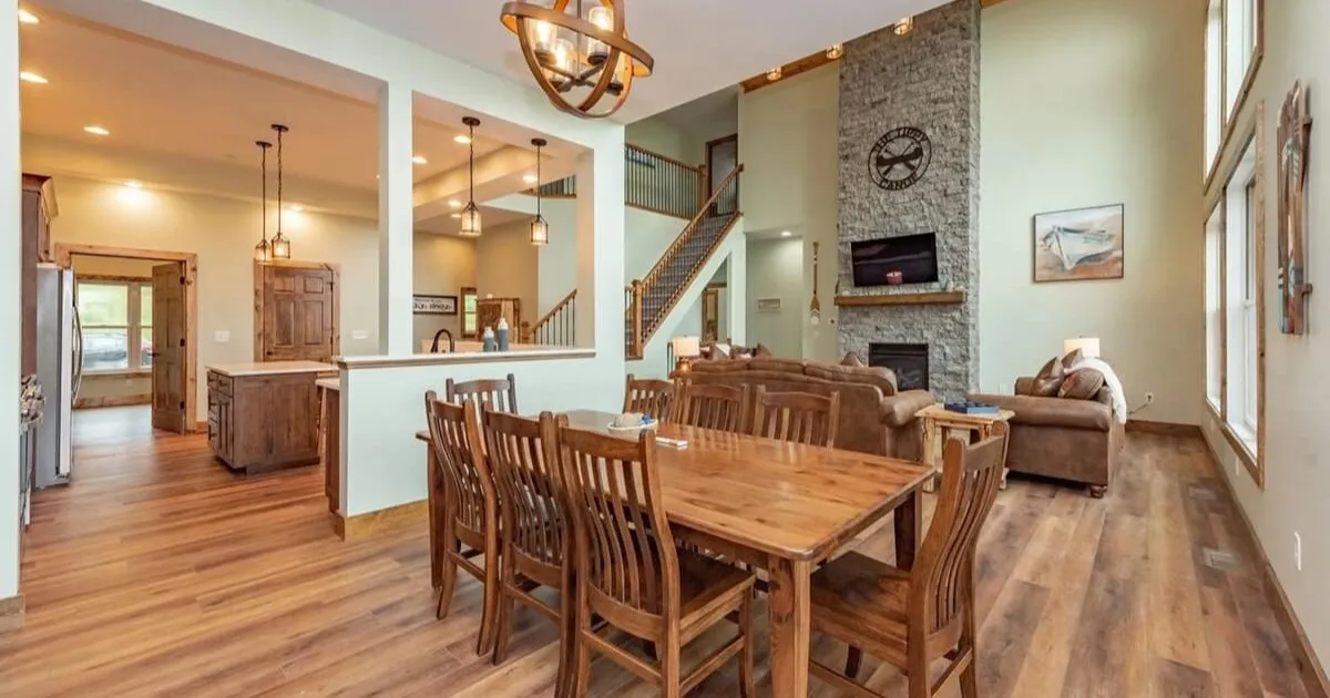 Open-concept dining and living area with a large wooden table, eight chairs, a kitchen island to the left, and a stone fireplace in the living room.