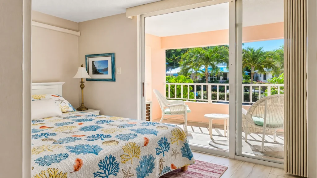 Hotel bedroom with a coral reef quilt, nightstand lamp, and a sliding door opening to a balcony with white chairs and palm trees outside.