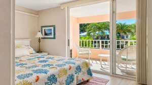 Hotel bedroom with a coral reef quilt, nightstand lamp, and a sliding door opening to a balcony with white chairs and palm trees outside.