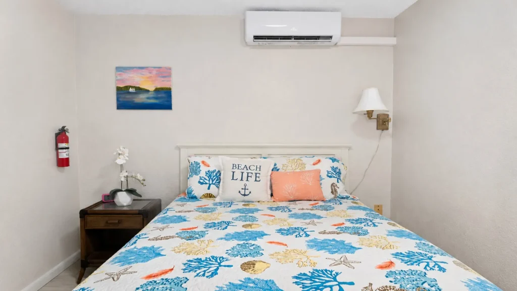 Coastal bedroom with a queen bed in a blue‑coral quilt, 'Beach Life' pillow, white headboard, wall lamp, and an AC unit above the bed.