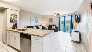 Open-concept kitchen and living area with a granite island, white cabinets, and stainless dishwasher, adjacent seating and a balcony view through blue curtains.