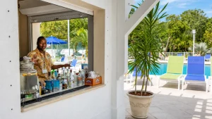 Poolside bar window with a bartender serving drinks; bottles and citrus jar on the counter, outdoor pool and colorful lounge chairs in the background.